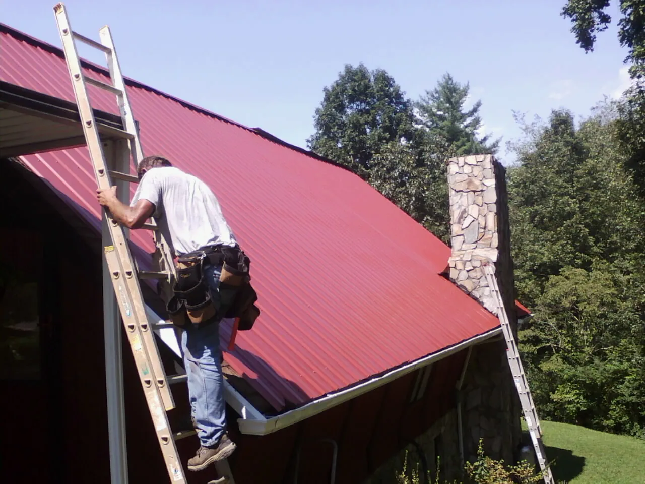 Roofer on a ladder beside a red metal roof and stone chimney — Soffit & Fascia Repair by Roofing Brothers in Prairie